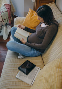 High angle view of woman reading book while sitting on sofa at home