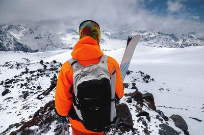 Portrait of a young male skier stands with his back with a backpack and skis in his hands against 