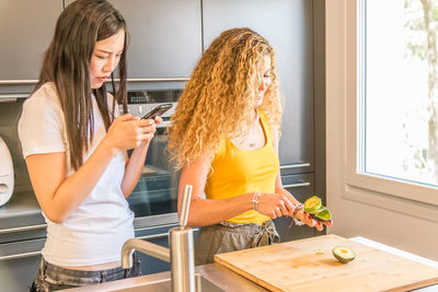 Woman using smart phone while friend cutting vegetables in kitchen