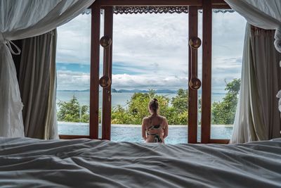 Rear view of shirtless man relaxing on bed against window