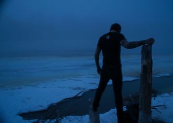 Rear view of man touching wooden post while standing at frozen river at dusk