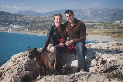 Portrait of couple with dog on rocky mountain against sky