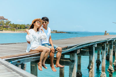 Full length of couple sitting on pier by sea against sky