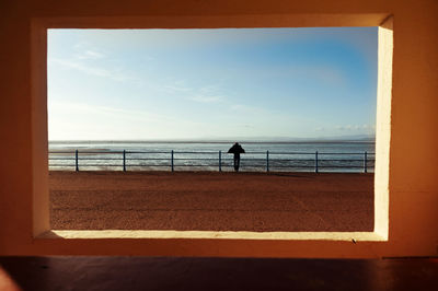 Rear view of man standing on promenade by sea seen through window