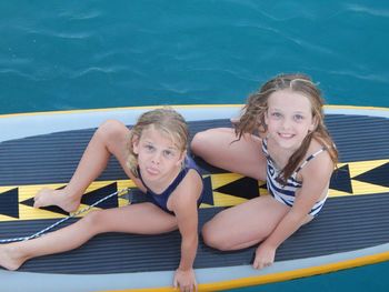 High angle portrait of cute sisters sitting on paddleboard in sea