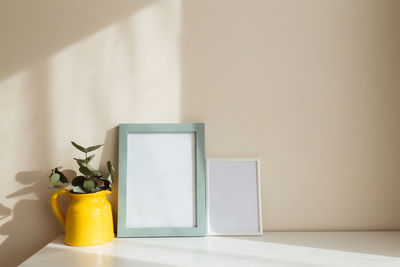 Empty white photo frames on the white table in the interior with beige walls near window.