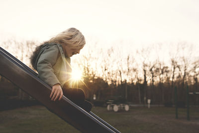 Side view of cheerful girl playing on slide at playground during sunset