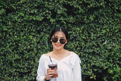 Portrait of smiling young woman holding drink glass against plants