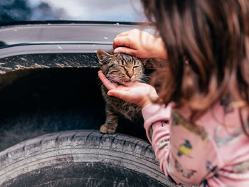 Crop girl in pajamas caressing adorable homeless tabby cat sitting on tire wheel of car