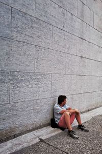 Side view of woman sitting on footpath against wall