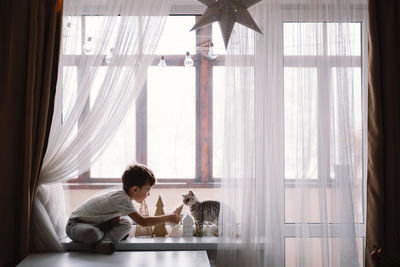 Cute boy playing with kitten sitting on the windowsill near the window. cozy home.