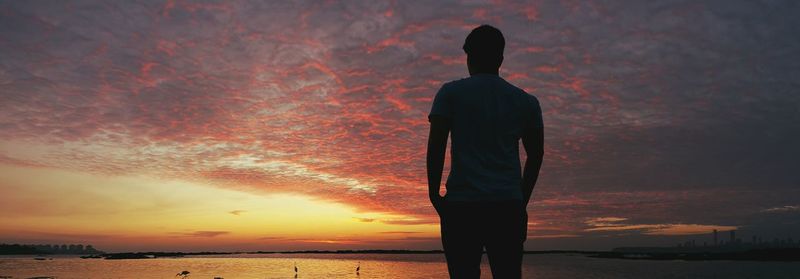 Rear view of man standing at beach against cloudy sky during sunset