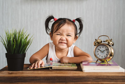 Cute girl reading book with alarm clock and potted plant on table at home