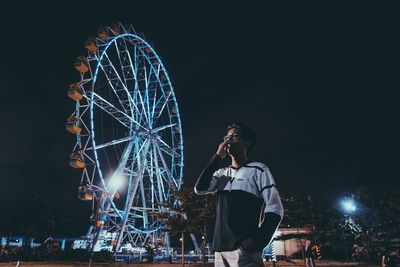 Low angle view of man standing by illuminated ferris wheel against sky at night