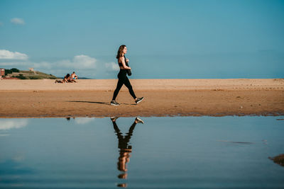 Side view of slim female in sportswear and with yoga mat walking along beach near river on sunny day
