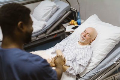 Male nurse holding hands of smiling senior patient lying on bed in hospital