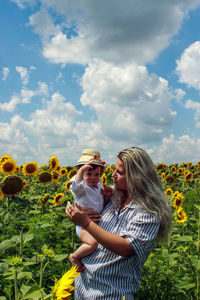 Mother holding son admist sunflower field against sky