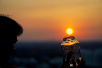 Close-up of man holding glass jar against sky during sunset