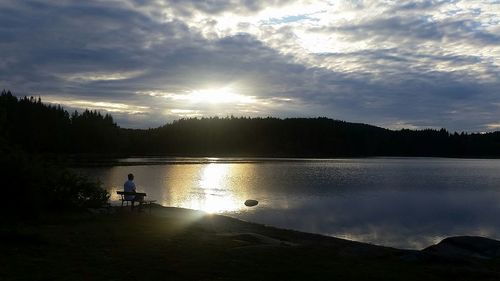 Silhouette man sitting by lake against sky during sunset