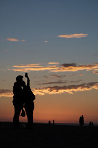 Silhouette couple taking selfie at beach against orange sky