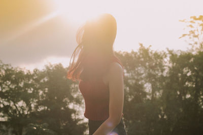 Side view of woman with tousled hair standing against sky during sunset