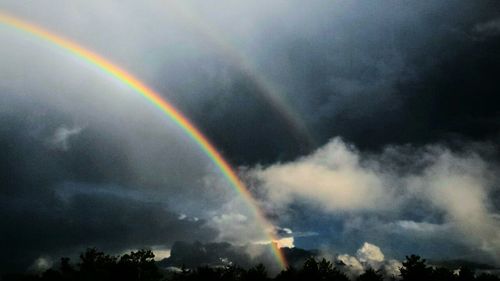 Low angle view of rainbow over trees against cloudy sky