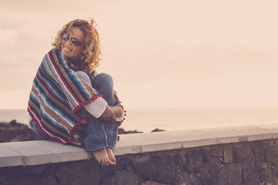 Woman sitting on retaining wall by sea against sky during sunset