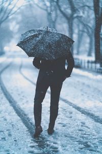 Rear view of man with umbrella walking on wet road during snowfall