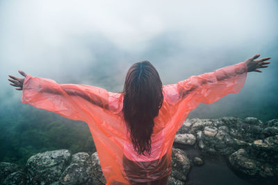 Rear view of woman with arms outstretched standing on cliff during foggy weather