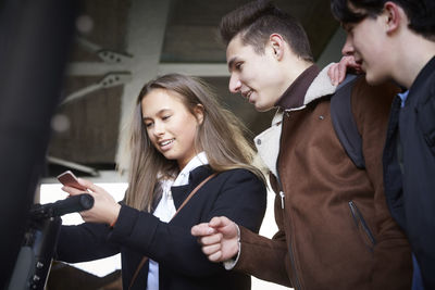 Teenage boys looking at female friend using mobile phone below bridge