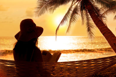 Rear view of woman reading book on hammock at beach during sunset