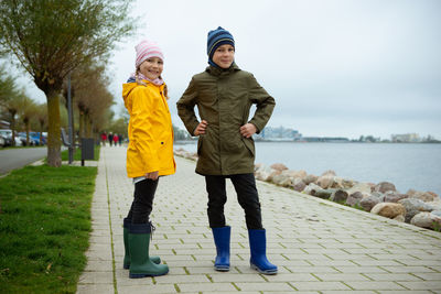 Full length portrait of girl standing with brother by sea on footpath