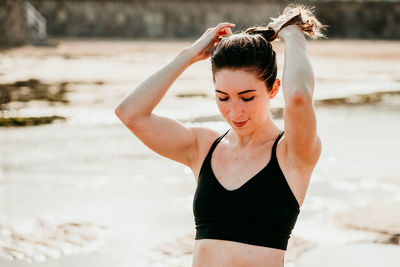 Peaceful female in sports bra standing on beach near river and making ponytail while preparing for yoga exercises