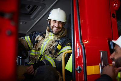 Smiling firefighter sitting in fire engine while talking to coworker