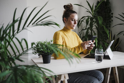 Young woman with digital tablet using mobile phone while sitting on table at home office