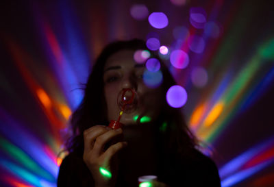 Portrait of woman blowing bubbles against illuminated lights