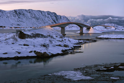 Bridge over river by snowcapped mountains against sky during winter
