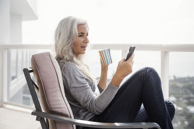 Side view of mature woman using smart phone while having coffee on balcony