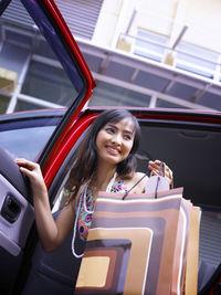 Portrait of smiling young woman holding shopping bags while getting out from car