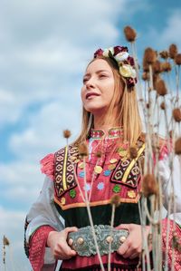 Beautiful young woman standing with flowers against sky