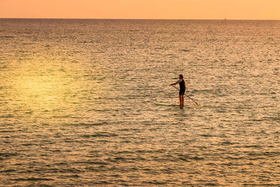  on beach during sunset. a man on a surfboard paddles standing by the sea at sunset