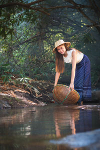 Portrait of smiling young woman holding basket by pond at forest