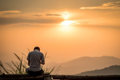 Rear view of man sitting on shore against orange sky