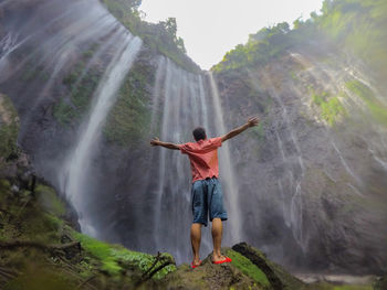 Rear view of man outstretching arms in front of waterfall
