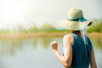 Rear view of woman wearing hat while standing against lake during sunny day