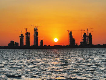 Silhouette buildings by sea against orange sky during sunset