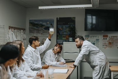 Teenage boy examining chemical beaker with friends and professor in science lab