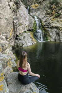Rear view of woman sitting on rock against waterfall