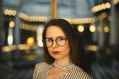 Portrait of young woman wearing eyeglasses against illuminated lights