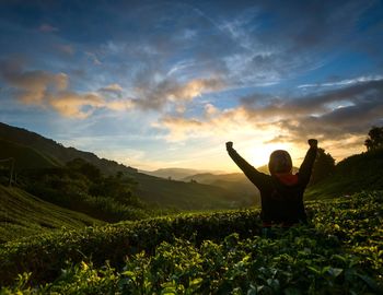 Woman with arms raised on field against sky during sunset